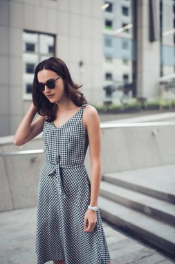 Çekici, kendine güvenen, güneş gözlüklü, zarif damalı elbisesi olan, tek başına merdivenlerde saçlarına dokunurken başka tarafa bakan ve düşünen çağdaş binaya bakan kendine güvenen bir kadın.