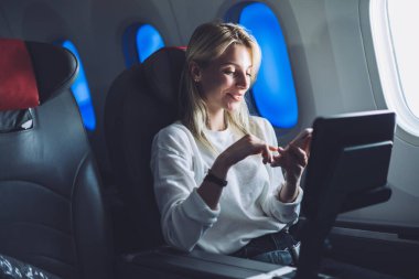 Modern uçakla rahat bir uçuşta cep telefonuyla sörf yaparken gülen rahat sarı saçlı kadın yolcu.  