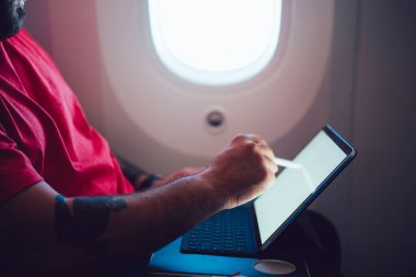 Kırmızı tişörtlü mahsul şahsın yan görünüşü uçuş sırasında bilgisayarda, iskele tarafındaki koltukta kablosuz olarak yaratıcı bir şekilde çizim yapıyor.