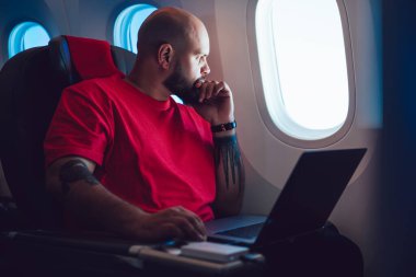 Düşünceli, sakallı, uzaktan kumandalı, kırmızı tişörtlü bir erkek kabin camından bakarken uçaktaki dizüstü bilgisayardaki projeyi analiz ediyor. 