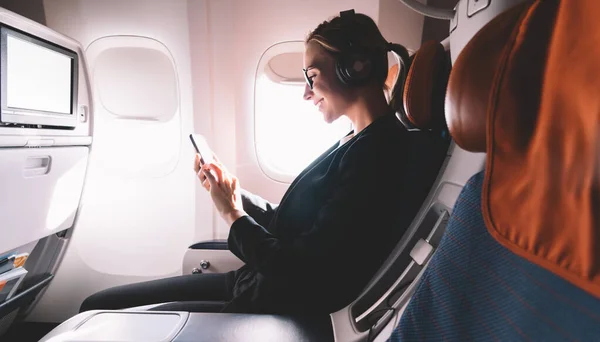 Female in formal wear listening audio record with info about intercontinental flights connected to wifi internet on mobile phone, cheerful woman with cellphone watching online movie and smiling