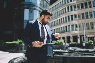 Resmi olarak sakallı genç adam kol saatini kontrol ediyor ve şaşırmış görünüyor. Çeşme ve binaların önünden geçerken kaşları yukarıda.