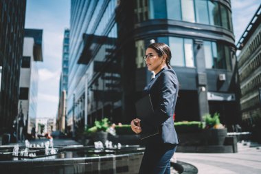 Resmi takım elbiseli, evrak işleriyle uğraşan, modern şehrin sokaklarında dikilip gözlerini çeviren bir kadınla buluşmayı bekleyen ciddi genç bir kadının yan görüntüsü.