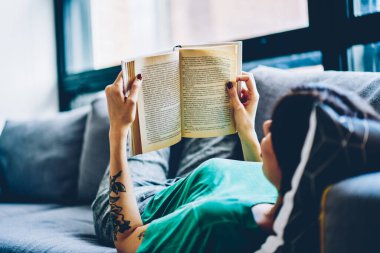 Back view of female holding open book on pages resting on comfortable sofa during weekend at home interior, 30s caucasian woman lying on couch reading interesting literature in living room on leisure