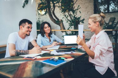Kafe terasında oturup uzaktan kumandalı bir iş yaparken neşeli iş arkadaşlarına gülümseyen ve belgeleri gösteren mutlu serbest yazar.