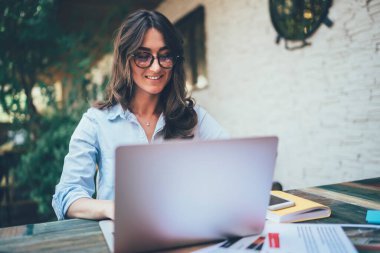 Gözlüklü, mavi tişörtlü, yeşil ağaç ve modern binanın beyaz duvarına karşı terasta otururken dizüstü bilgisayarla etkileşen oldukça düşünceli yetişkin bir kadının alçak açısı.