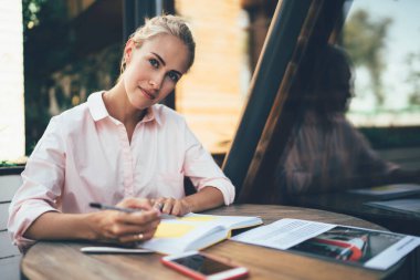 Düşünceli bakışlı genç bir kadın kafenin terasındaki masada oturuyor ve not defterine yazmak için yazı yazıyor.
