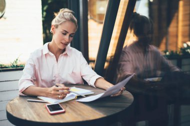 Kafe terasında, masada oturan, elinde defterle dikkatle reklam broşürü okuyan, iş teklifiyle ilgilenen genç bir kadın. 