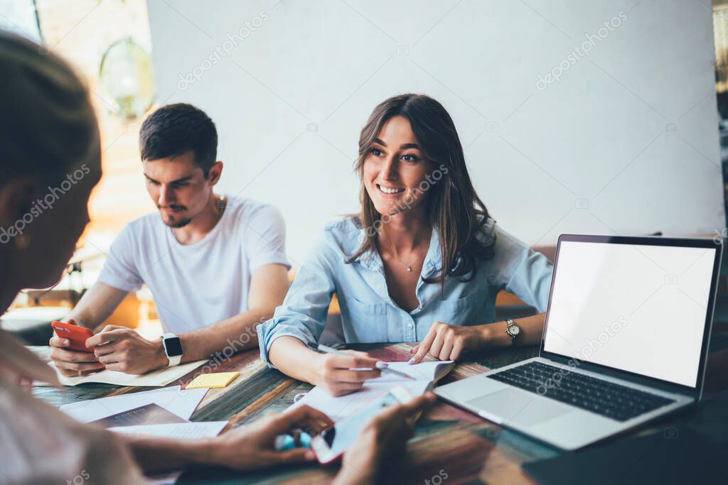 Ethnic woman with notebook talking to female teammate with smartphone and laptop with blank screen while sitting at table with male coworker surfing Internet