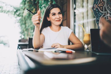 Elinde meyve suyu dolu not defteri ve kalemle kafede oturan ve dizüstü bilgisayarı olan tanınmayan biriyle konuşan pozitif destekleyici genç bir kadın.