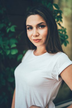 Beyaz gömlekli, kendine güvenen genç bir kadın. Arka planda yeşil ağaçlara karşı el pençe divan duruyor.