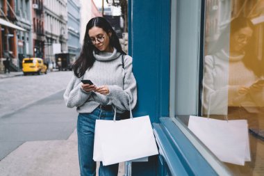 Güzel, ince, çekici, gözlüklü, alışveriş torbalı kot ve kazaklı bir kadın. Şehir caddesinde faiziyle akıllı telefon kullanıyor.