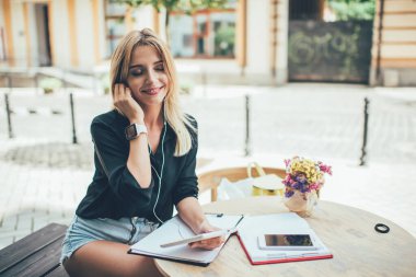 Elektronik kulaklıklı mutlu beyaz öğrenci kaldırım kafeteryasında sınav hazırlıkları sırasında pozitif müzik listesi dinliyor kulaklıklarıyla kulaklık takan kaygısız bayan müzik aşığı açık havada dokunmatik ped kullanıyor.