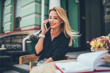 Neşeli kadın gazeteci, eğitim kitabıyla kaldırım kafede oturuyor ve arkadaşıyla uluslararası sohbet yapmak için modern cep telefonu cihazında gezinen internet bağlantısını kullanıyor.