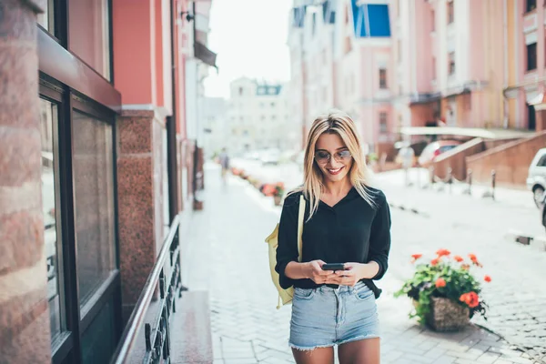 夏休みに街の通りを歩きながらGpsトラッキングのための現代のスマートフォンガジェットで4Gの無線を使用して陽気な女性観光客 スタイリッシュなサングラスブラウジングウェブサイトで幸せな女性 — ストック写真