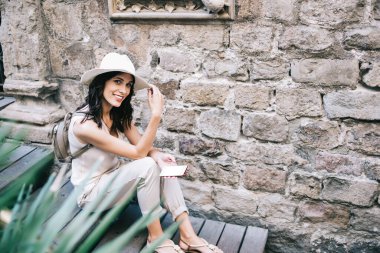 Yüksek açılı, gündelik giysiler içindeki çekici bir kadın ahşap merdivenlerde otururken kameraya bakıyor ve şapkaya dokunuyor ve ortaçağ taş binasına karşı akıllı telefonla iletişim kuruyor.