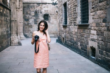 Eski turizm caddesinde kaldırımda dikilirken gülümseyen ve saçlarına dokunan fotoğraf makineli arkadaş canlısı genç bayan.
