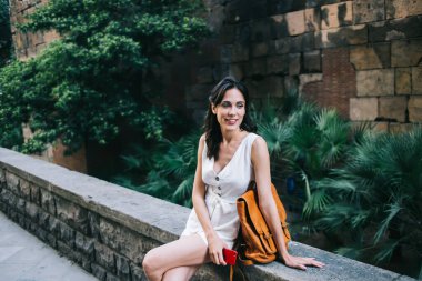 Kısa beyaz kolsuz, büyük turuncu çantalı, elinde kırmızı cep telefonuyla oturan gülümseyen romantik kadın.