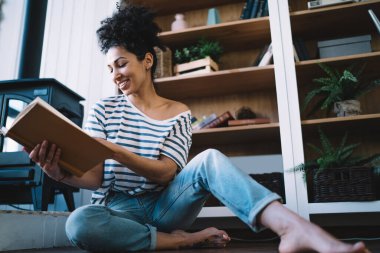 Sade giyinmiş, şöminenin yanında kıvırcık saçlı, kitaplıkların yanında oturmuş, çağdaş düzlükte roman okuyan, huzurlu, etnik bir kadın.
