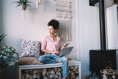 Dekoratif bankta bacakları çapraz oturan düşünceli, düşünceli bir etnik kadın. Tahtaya yer var ve cep telefonuyla dizüstü bilgisayarda yazıyor. 