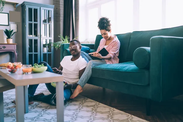 Happy adult African American man in casual clothes looking at camera and interacting with smartphone while sitting on floor at table near pleased ethnic woman reading book on sofa in light modern apartment