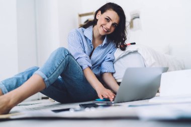 Modern odada çıplak ayakla grafikler ve kağıtlarla yerde otururken günlük kıyafetleriyle bilgisayarı yazan neşeli bir kadının yan görüntüsü.