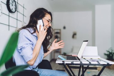 Kahkaha atan kahverengi saçlı, gözlüklü, cep telefonuyla konuşan ve evde dizüstü bilgisayarla çalışan bir kadının yan görüntüsü.