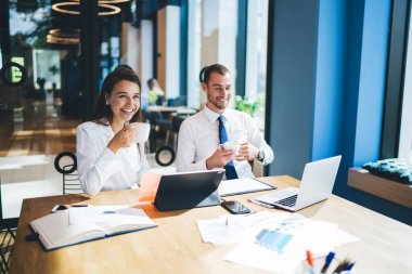 Neşeli erkek ve kadın girişimciler iş için canlı iletişimin keyfini çıkarıyorlar. Beyin fırtınası yapıyorlar. Masanın içinde oturmuş gülümsüyorlar ve kahve molası veriyorlar.