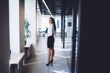 Resmi giysiler içinde, ofis koridorunda duran ve kameraya gülümseyen başarılı yönetici kadın, resmi kıyafetli neşeli bayan girişimcinin tam boy portresi.