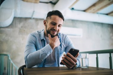 Düşünceli hippi adam cep telefonundan mesaj okuyor sosyal ağlarda sohbet ediyor, kahve molasında gülümsüyor, milenyum boyunca akıllı telefondan video izliyor ve uygulamadan keyif alıyor ve 4G bağlantısı kuruyor.