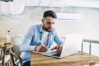 Kafkasyalı 20 'li yaşlarda erkek öğrenci ödevlerini not defterinde yazmaya ve kafenin içindeki kafeteryada kullanmaya odaklanmış. Düşünceli adam bilgisayardaki internet sayfasında bilgi arıyor.