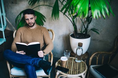 Kendine güvenen 20 'li yaşlarda bir erkek, kafenin içinde oturmuş, yeni başlayanlar için bilgi topluyor. Düşünceli hippi adam ilginç bir kitaptan bilgi alıyor.