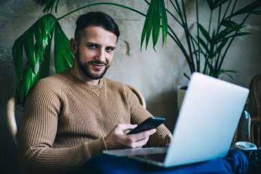 Uzak iş için dijital cihazlar ve internet bağlantısı kullanarak kafenin içinde oturan milenyum erkek serbest yazarının portresi, pozitif hipster erkek blogcu sosyal ağlarda medyayı paylaşıyor. Kameraya bakın.
