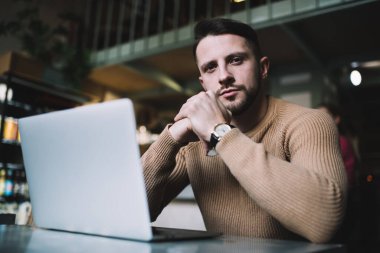Laptop bilgisayarının yanındaki kafede oturan sıradan giyinen yakışıklı iş adamının yarım boy portresi, araştırma ve planlama projesine odaklanmış kameraya bakan ciddi bir erkek girişimci.