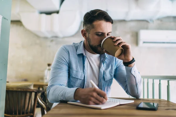 Young skilled 20s male blogger writing publication and articles in notepad while drinking caffeine beverage for productive remote job, hipster guy enjoying coffee while learning and doing hometask