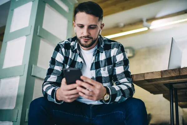 Kulaklıklı genç beyaz erkek cep telefonundan 4G bağlantısı ve bluetooth üzerinden video izlemekten zevk alıyor. Düşünceli hipster Meloman, cep telefonuna müzik listesi müziği dinlediği için uygulama yüklüyor.