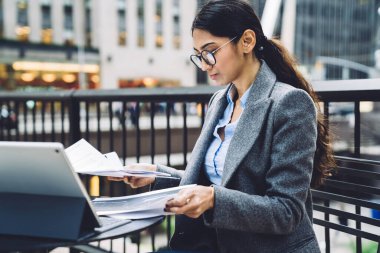 Zarif kıyafetli ve gözlüklü zeki etnik iş kadını New York şehrinin terasındaki tableti incelerken açık havada masalarda oturup belgeleri kontrol ederken görülüyor. 