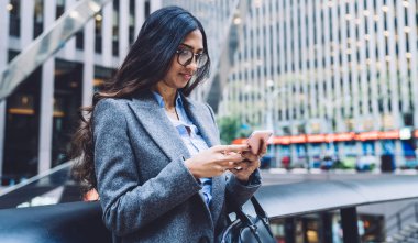Gözlüklü, modaya uygun ceketli, deri çantalı ve şehrin arka planında duran telefonu kullanan kendinden emin genç bir bayan.