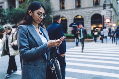 Düşünceli, zarif, zarif, esmer, gözlüklü, siyah çantalı, gri ceketli bir kadın. New York 'ta kalabalık bir sokakta yaya geçidinde, bulanık arka planda akıllı telefona dokunuyor.