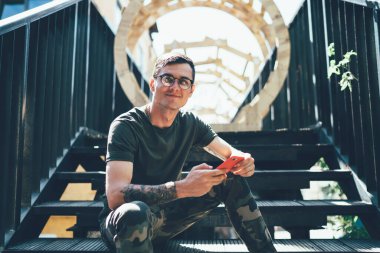 Yeni nesil gözlüklü yakışıklı hippinin portresi göz koruması sağlamak için kameraya bakarken akıllı telefon cihazına uygulama takmayı bekleyen, gençliğin tadını çıkaran genç bir adam.