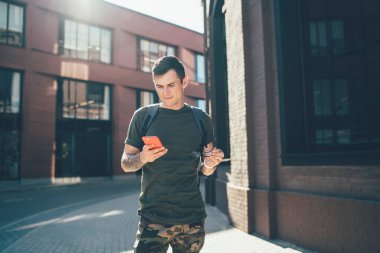 Sırt çantalı, gözlüklü ve ekranda dikilip güneşli bir günde bulanık modern binalara karşı akıllı telefon kullanan düşünceli bir adamdan.