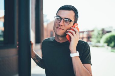 Günlük kıyafetleri ve modaya uygun gözlükleri olan mutlu genç bir erkek öğrenci yığını tutarken ve şehir caddesine bakarken cep telefonundan arıyor. 