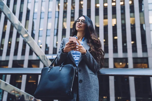 ニューヨークを背景に電話で革のバッグをチャットし 離れて見て眼鏡やコートを保持現代の民族ビジネス女性を自信を持って — ストック写真