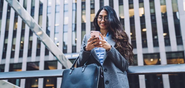 古典的なコートやシャツの眼鏡で若い民族の女性は 市内の笑顔を背景に電話で革のバッグメッセージングと — ストック写真