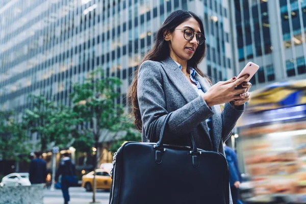 眼鏡や革のバッグを運ぶ流行のコートで現代の民族の女性とニューヨーク市の背景に携帯電話の歩行を使用して — ストック写真