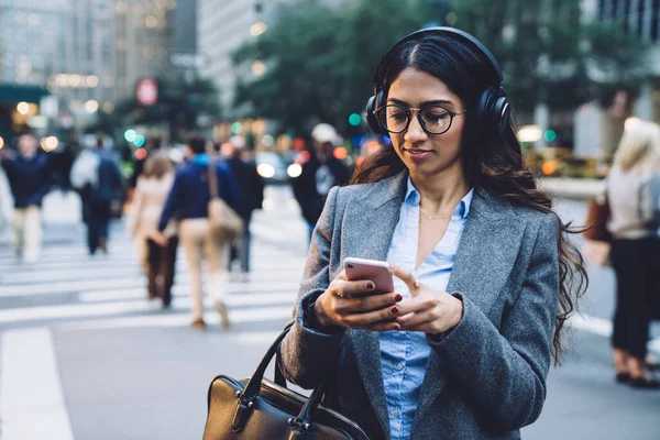 Modern young woman in formal wear glasses using headphones and smartphone for listening to music while walking in New York