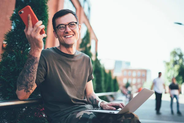 Happy male student with cellular phone in hand laughing from funny joke read on website with comedy content while spending time in campus with laptop computer for e learning, concept of generation