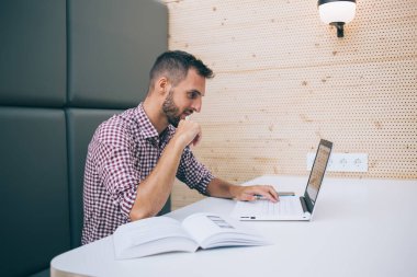 Modern kafede masa başında, dizüstü bilgisayarla uzaktan kumandalı proje üzerinde çalışan, gündelik giysiler içinde konsantre genç serbest çalışan biri.