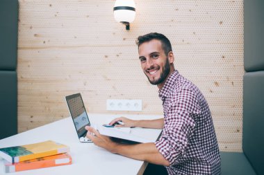 Modern işyerindeki masada oturup kameraya bakarak dizüstü bilgisayar kullanarak evrak işleriyle uğraşan serbest giyinen etnik ve güler yüzlü bir serbest yazar.