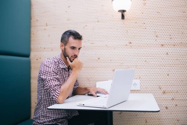 Ciddi odaklı, sakallı, sıradan tişörtlü, çenesine dokunan diz üstü bilgisayarlı mavi koltukta oturan ve duvarında lamba olan hippi.